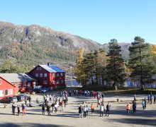 Nesholmen leirstad med born og unge utandørs — raud bygning og fjell i bakgrunnen